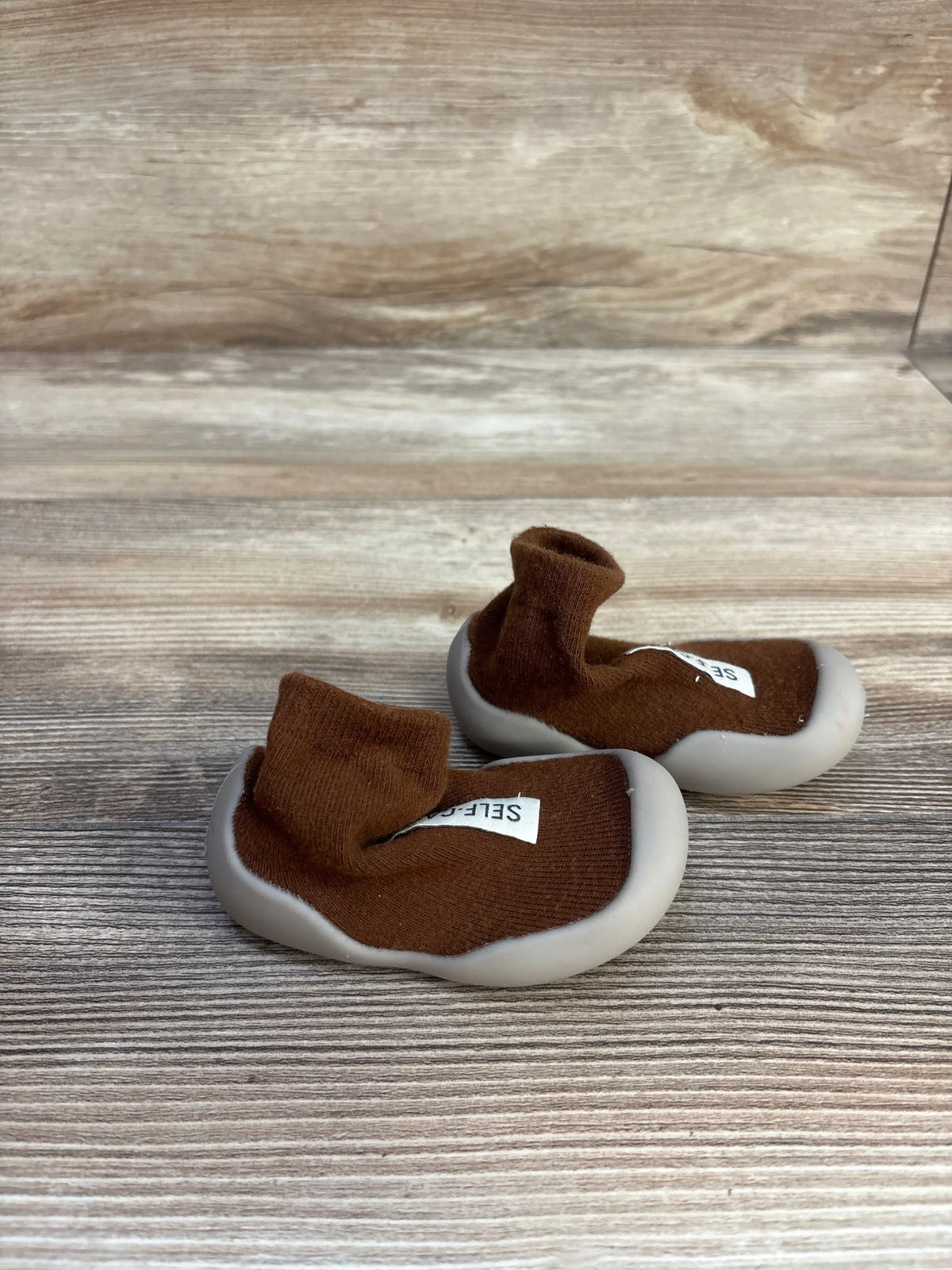 Angled view of Tiny Explorings Self-Care Sock Shoes in brown, size 2/3C, showcasing sock-style uppers and light gray soles on a wood-look floor