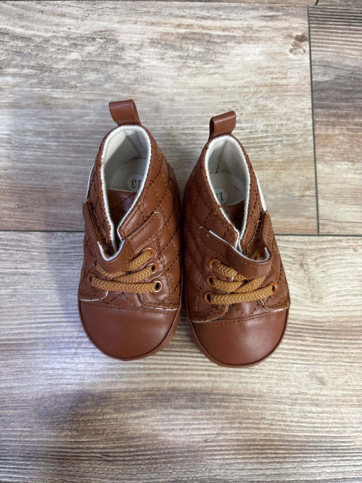 Front view of brown faux leather crib shoes showcasing quilted uppers and lace-up fronts on a wood-grain surface
