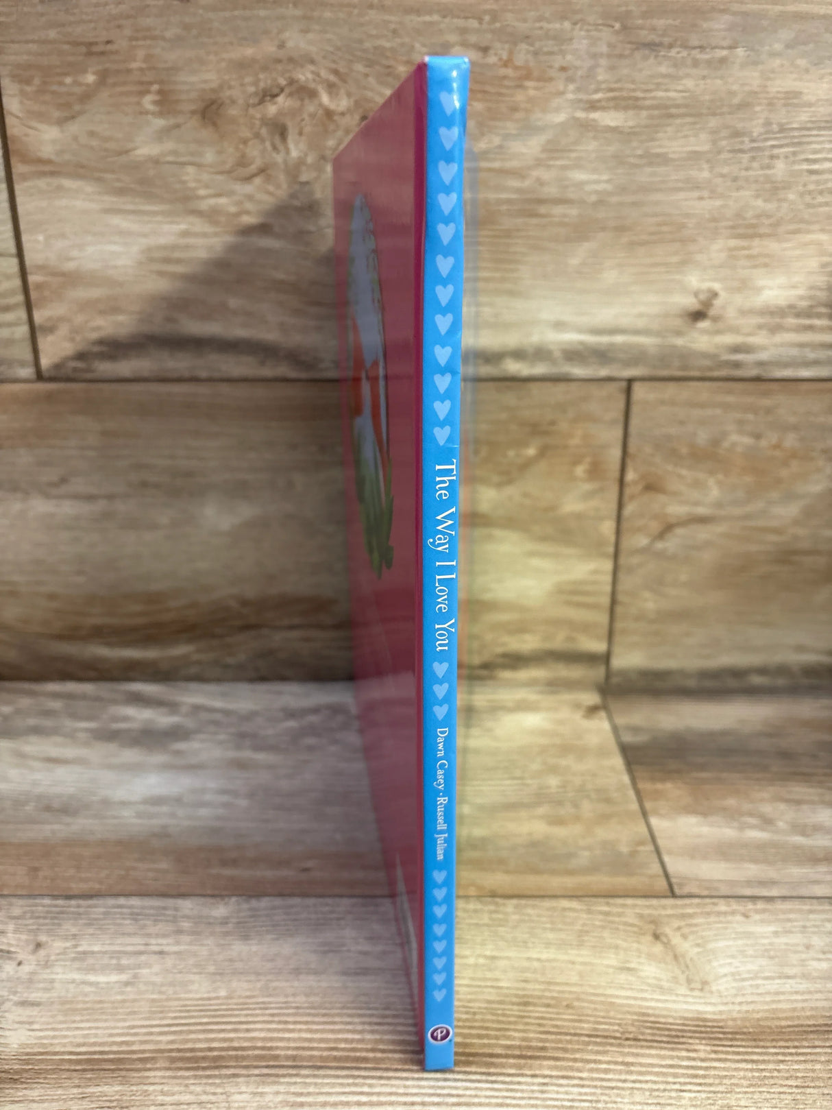 Side view of The Way I Love You hardcover book showcasing blue heart-patterned spine and vertical title with wooden shelf background