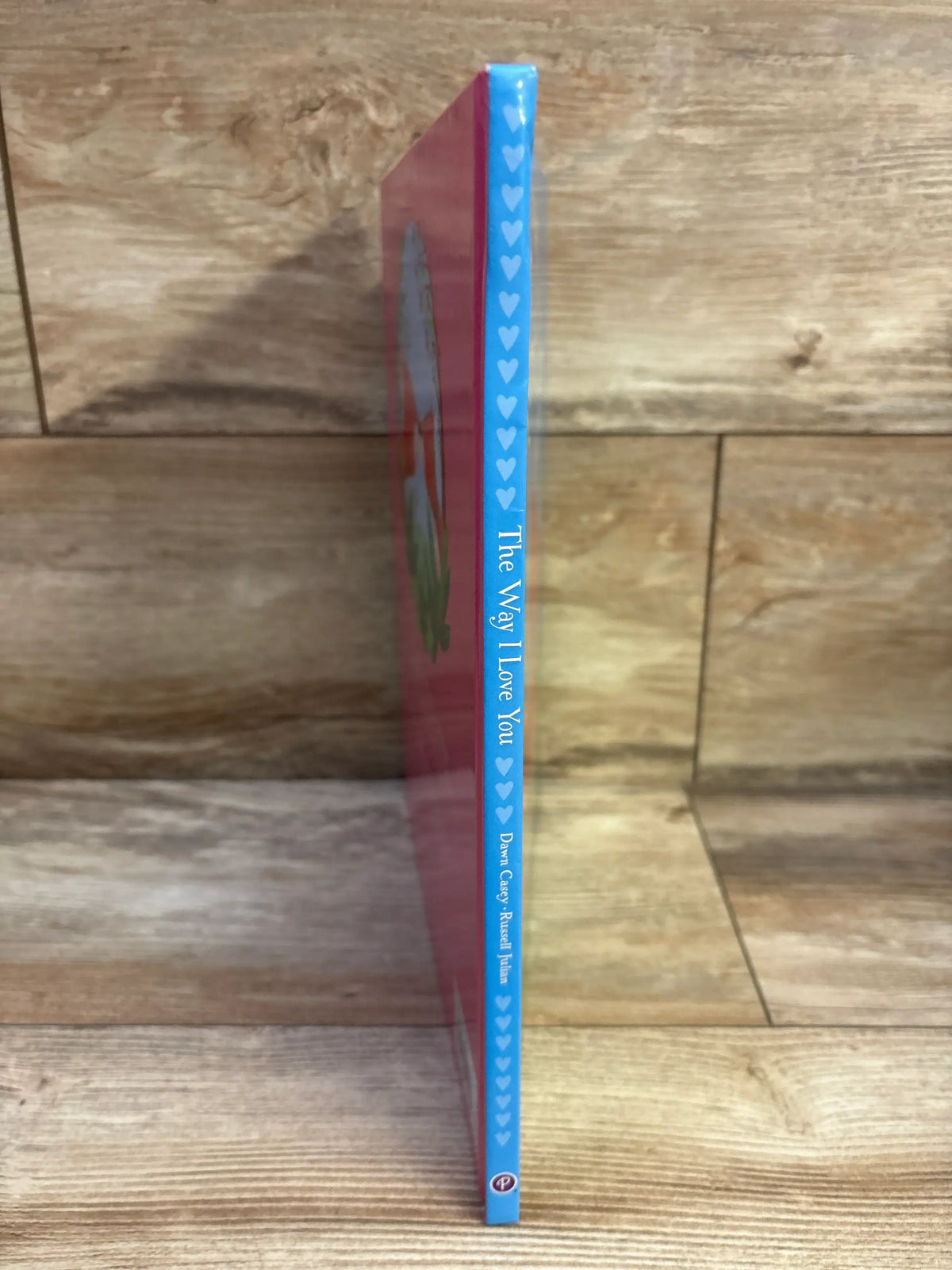 Side view of The Way I Love You hardcover book showcasing blue heart-patterned spine and vertical title with wooden shelf background