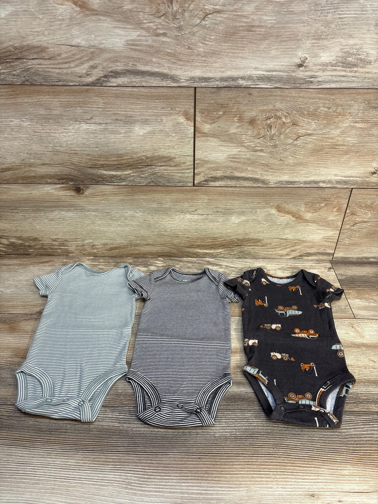 Three baby onesies: striped, navy striped, and black construction vehicle print on wood floor