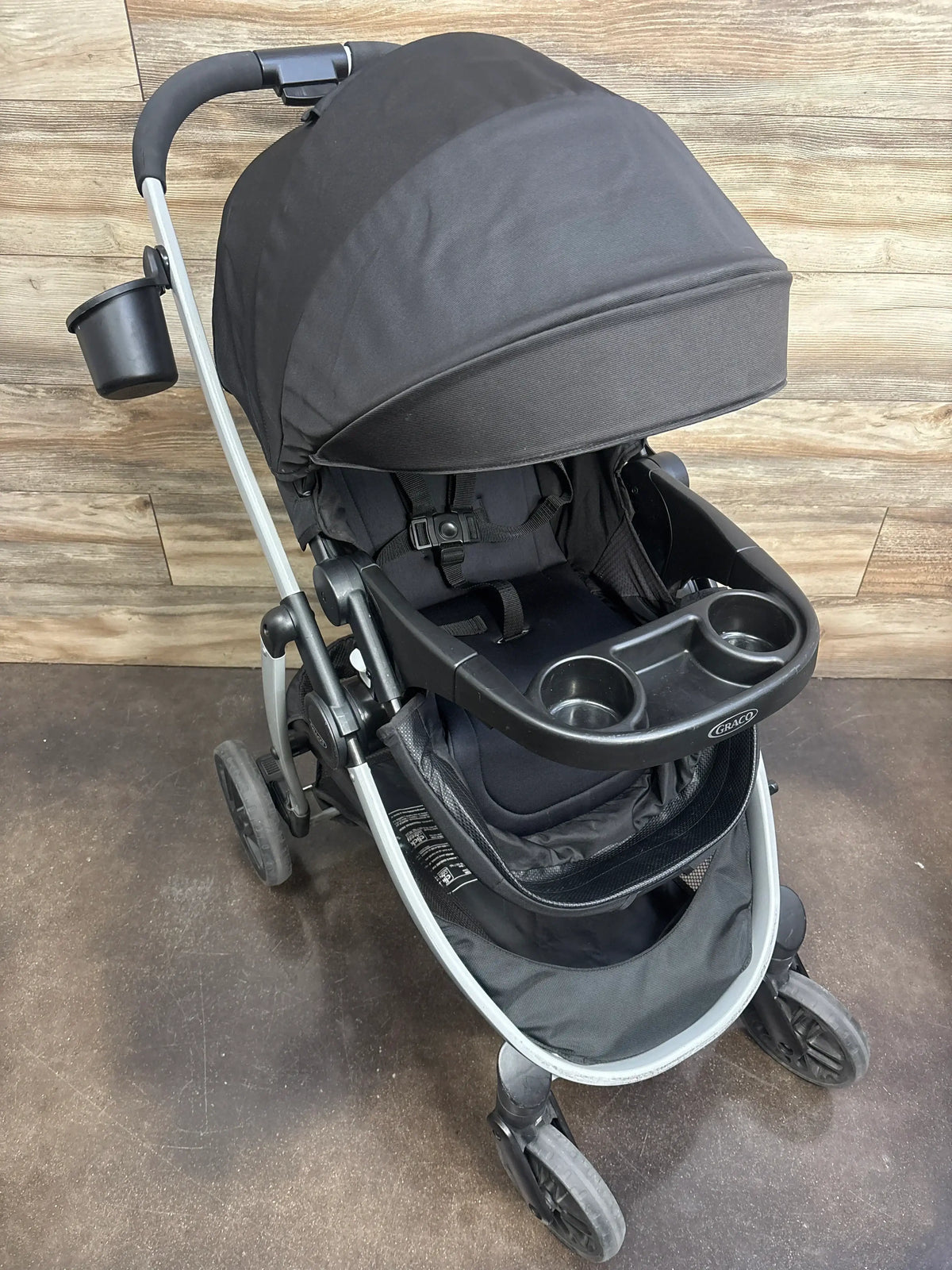 Front three-quarter view of black Graco Modes Pramette Stroller showcasing covered seat, harness, cup holder tray, and storage basket indoors