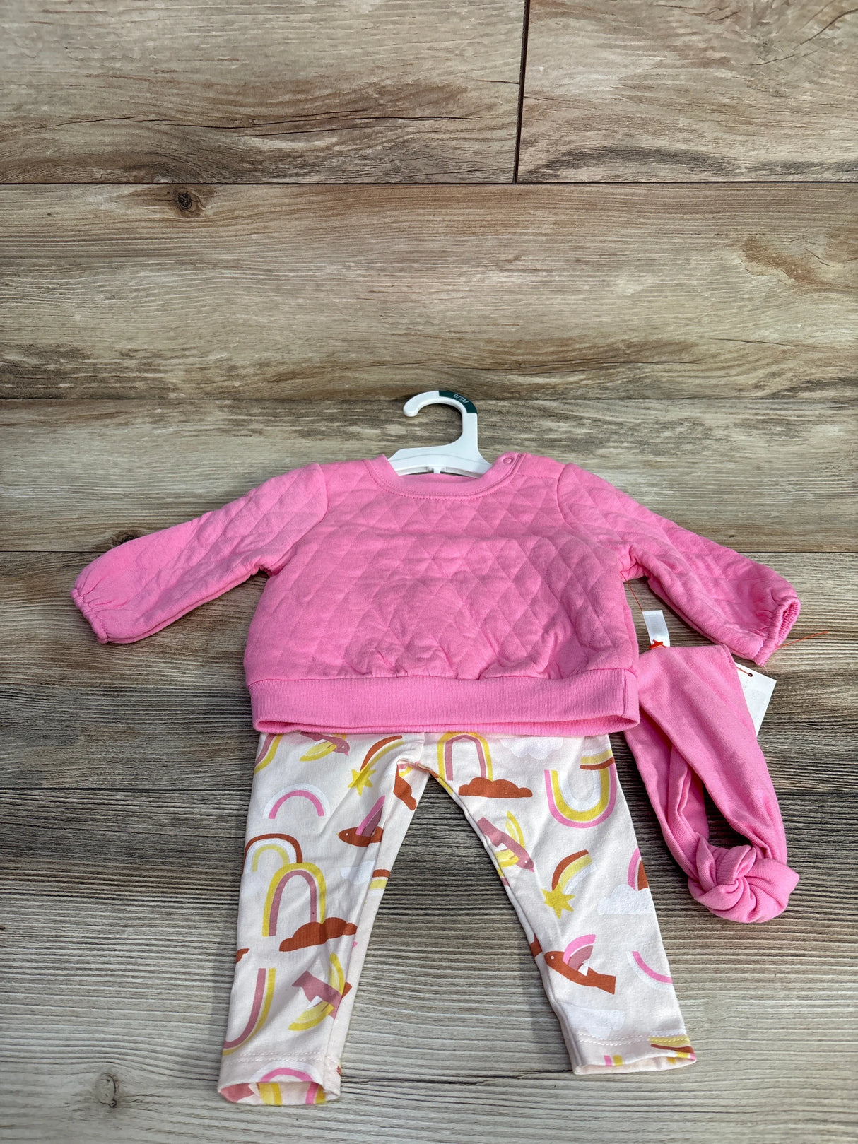 Pink quilted baby sweater, white leggings with colorful rainbows and birds, and pink knotted hat on wooden floor