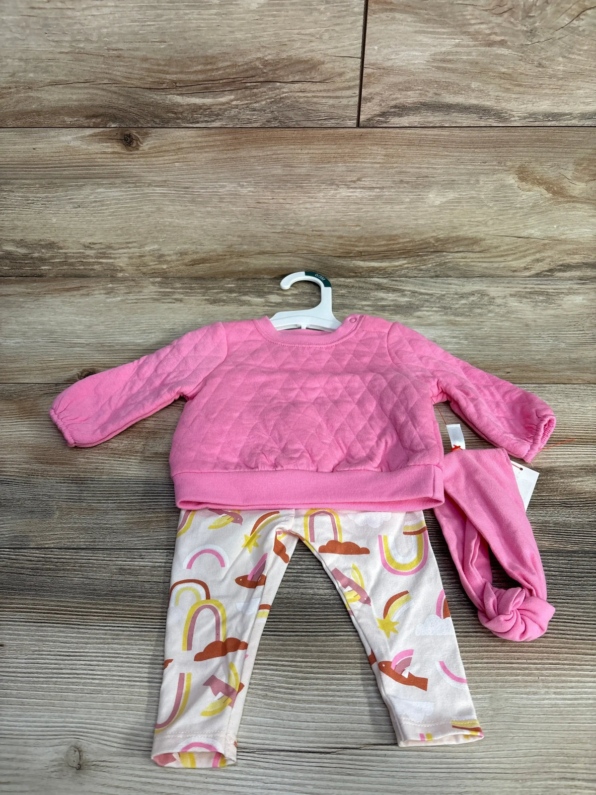 Flat lay of newborn 3-piece quilted pink sweatshirt and pants set with rainbow-and-bird leggings and pink knotted hat on wooden floor