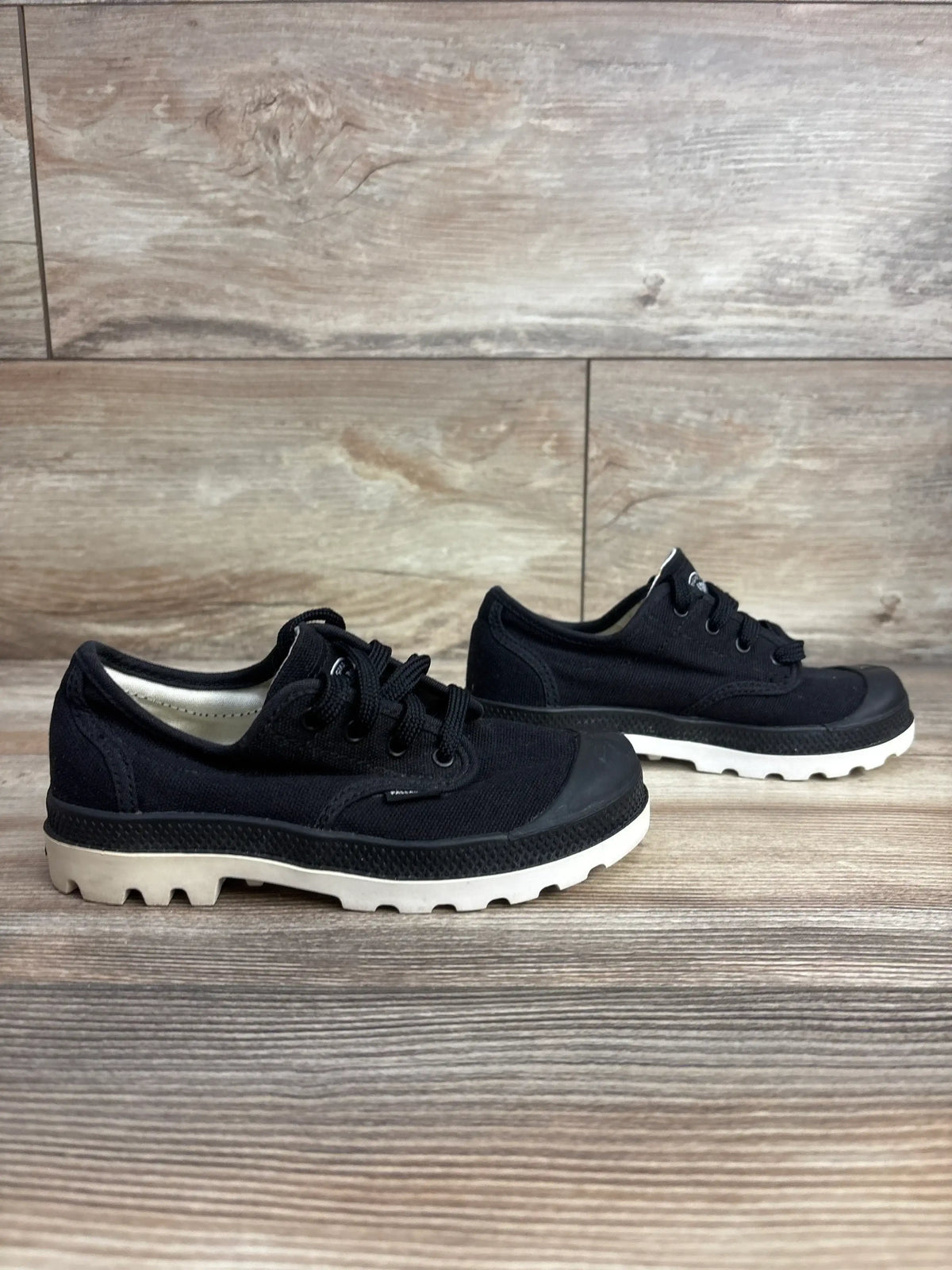 Side view of Palladium Pampa Oxford kids sneakers in black showing canvas upper and white lugged rubber sole on wooden floor