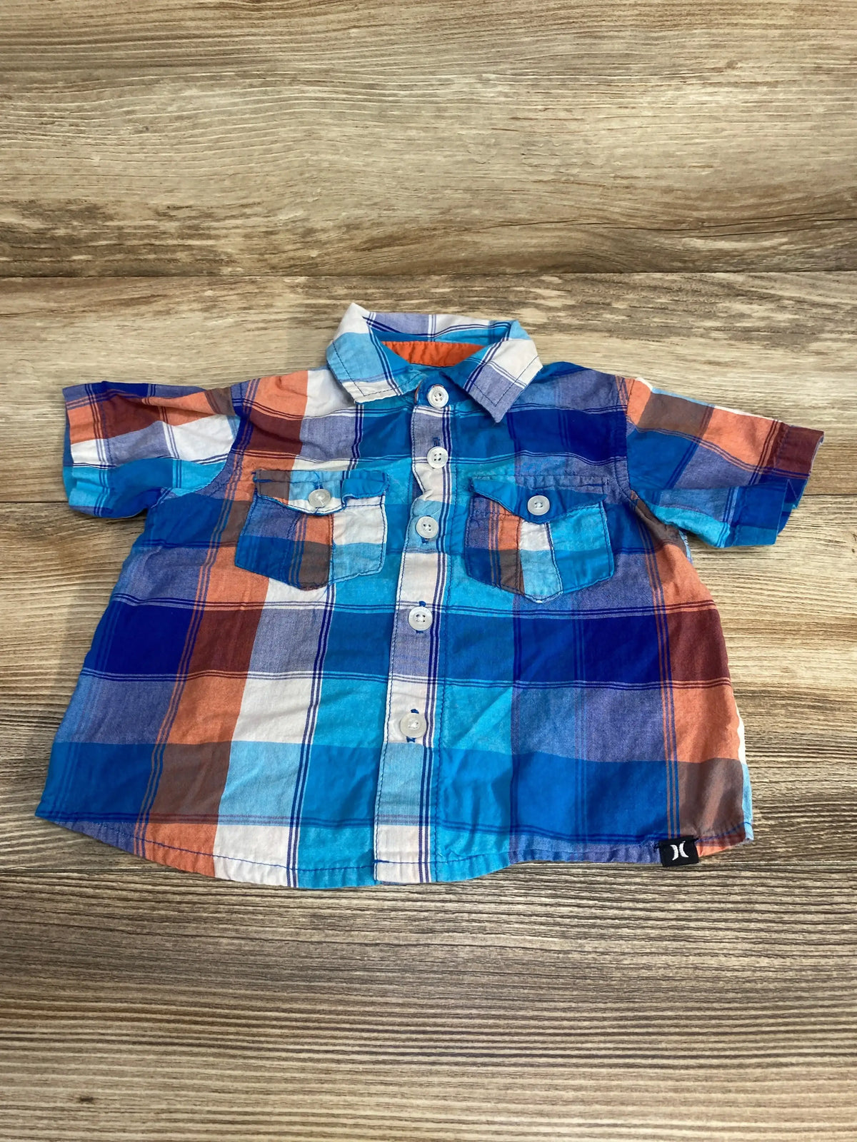 Front view of toddler Hurley plaid button up shirt in blue, orange, and white with chest pockets on wood background