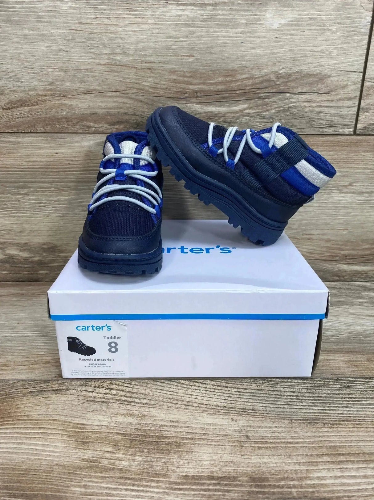 Side view of Carter’s toddler Fallon boots, navy blue, size 8, showing lug soles and lace-up on box on wood backdrop