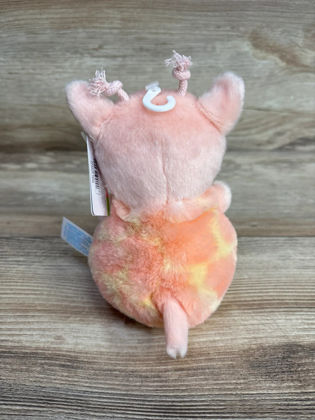 Back view of ebba Baby Giraffe Plush showing pink and orange tie dye body, braided ossicones and small tail on wooden background