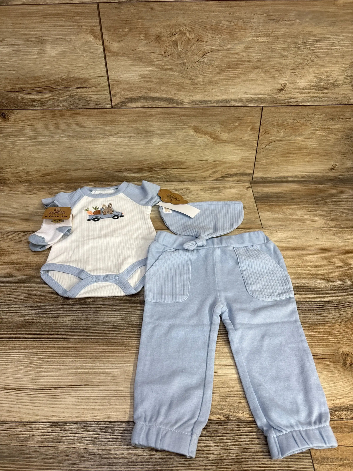 Flat lay of light blue baby bodysuit and pull on pants set showing bunny in a truck embroidery and ribbed pocket details with matching hat and socks on wood background