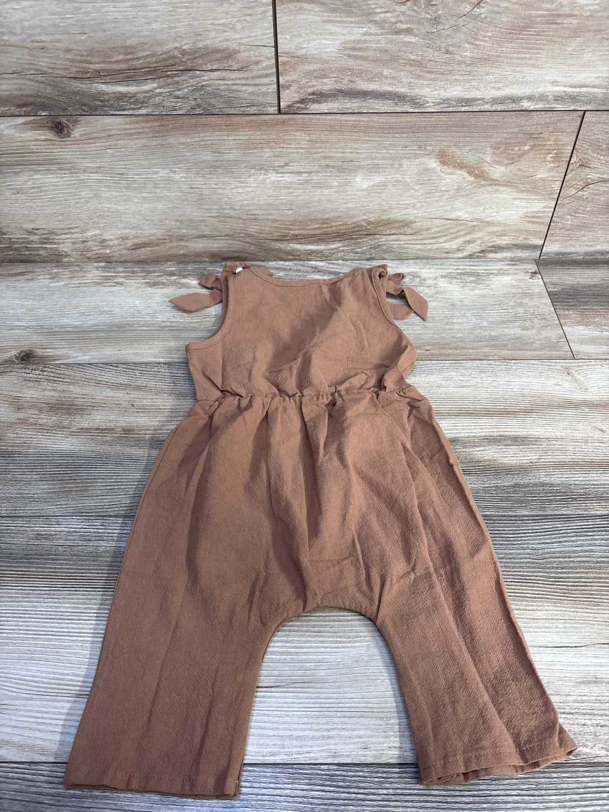 Flat lay of brown sleeveless baby jumpsuit with tied shoulder bows and gathered waist on wood background