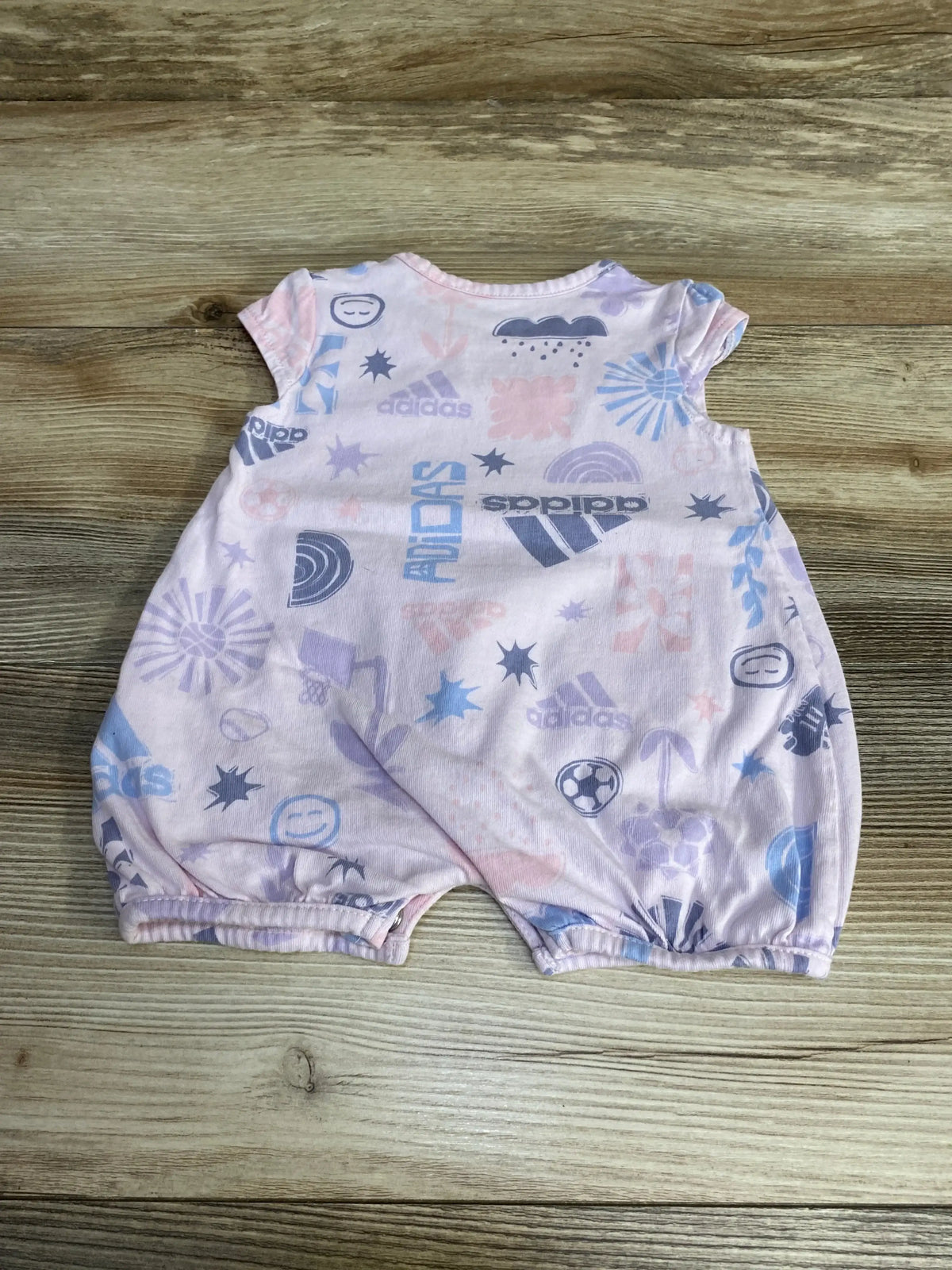Flat lay of pink Adidas shortie romper (3M) showing all-over logos and pastel sports icons on wood floor