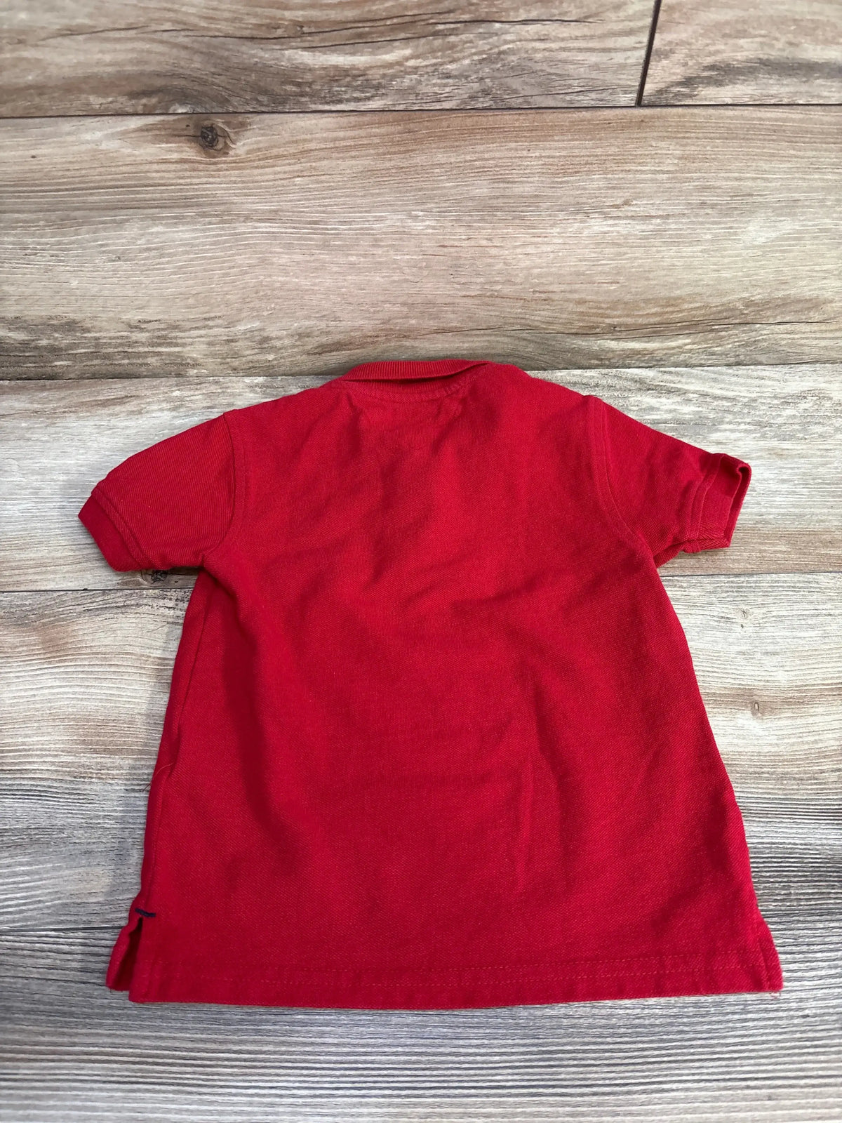 Back view of red Tommy Hilfiger 2T toddler polo shirt showing short sleeves, ribbed collar, and side vents on wood background