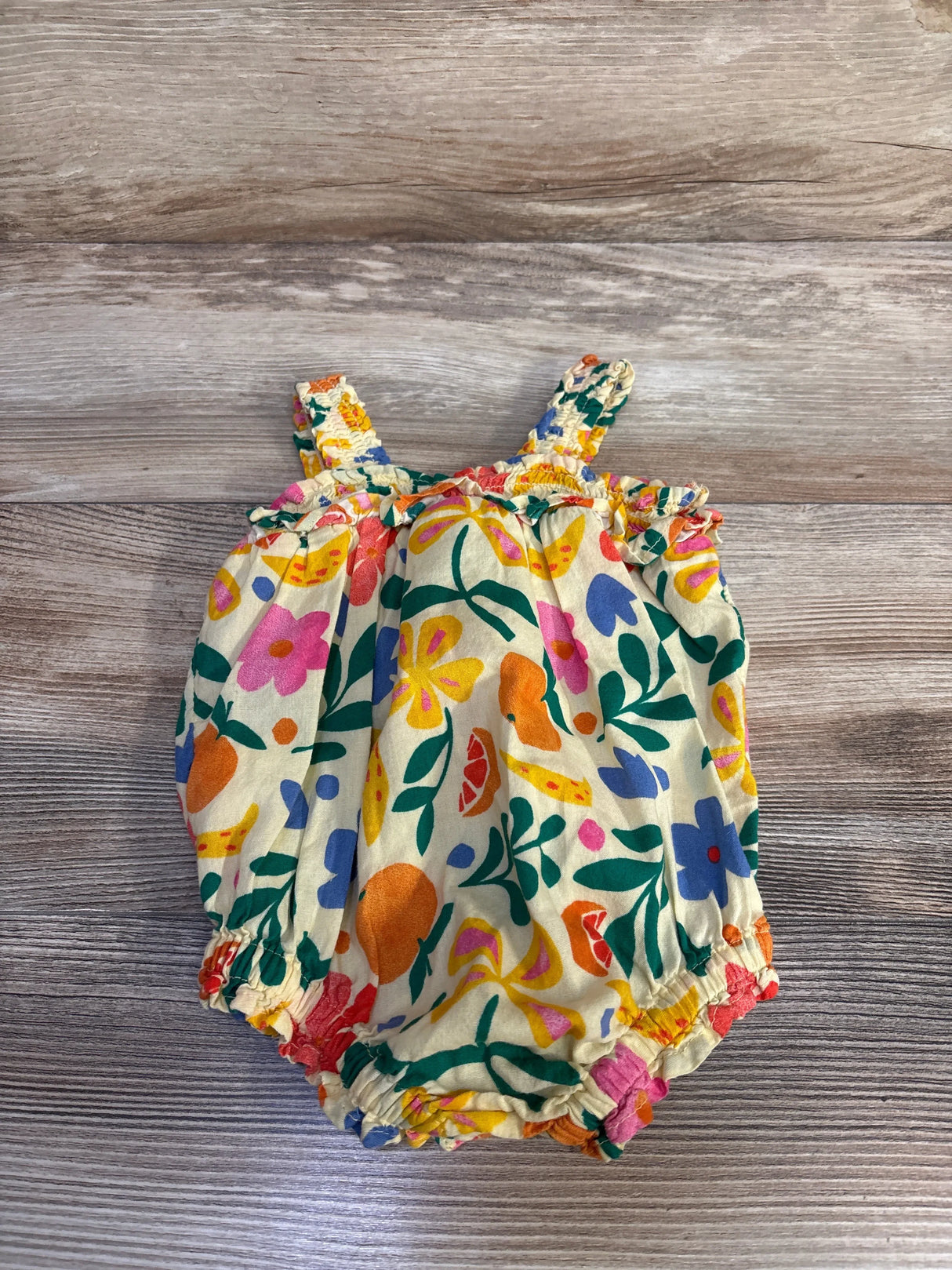 Flat lay of yellow smocked bubble romper with multicolor floral print, ruffle trim, elastic leg openings, on wood background