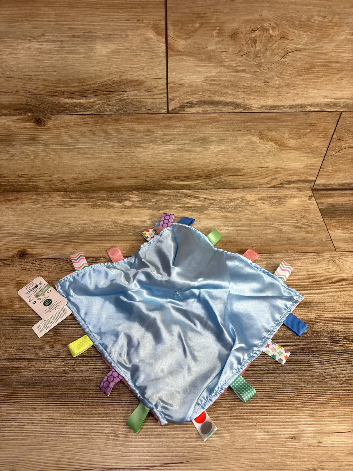 Flat lay of Taggies Camilla caterpillar lovey blanket showing light blue satin and multicolored ribbon tags on wood floor