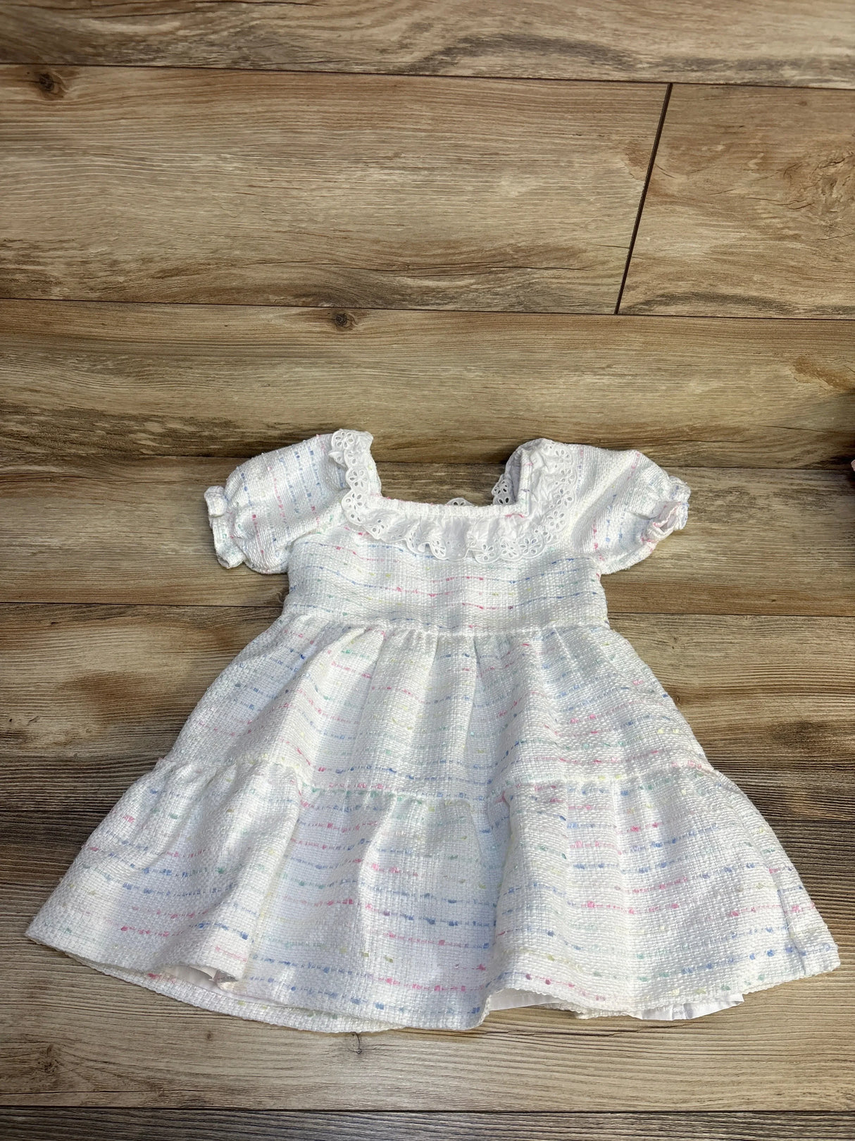 White baby dress with pastel stripes, lace trim, and puffed sleeves on wood floor