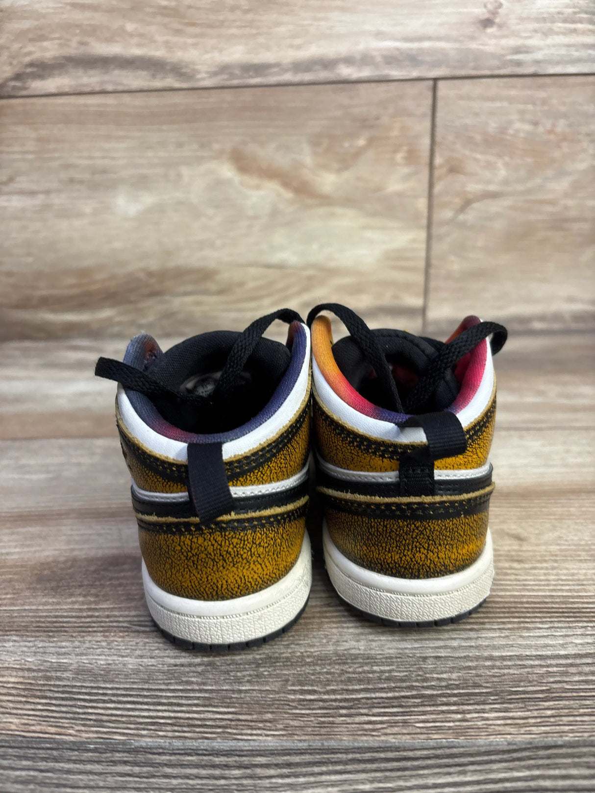 Rear view of toddler Air Jordan 1 Mid SE Wear-Away Taxi sneakers with cracked yellow finish and rainbow lining on wood