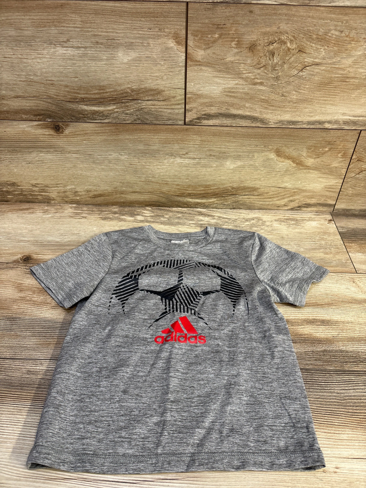 Flat-lay front of toddler Adidas grey short-sleeve soccer shirt showing black soccer-ball graphic and red Adidas logo on wood background