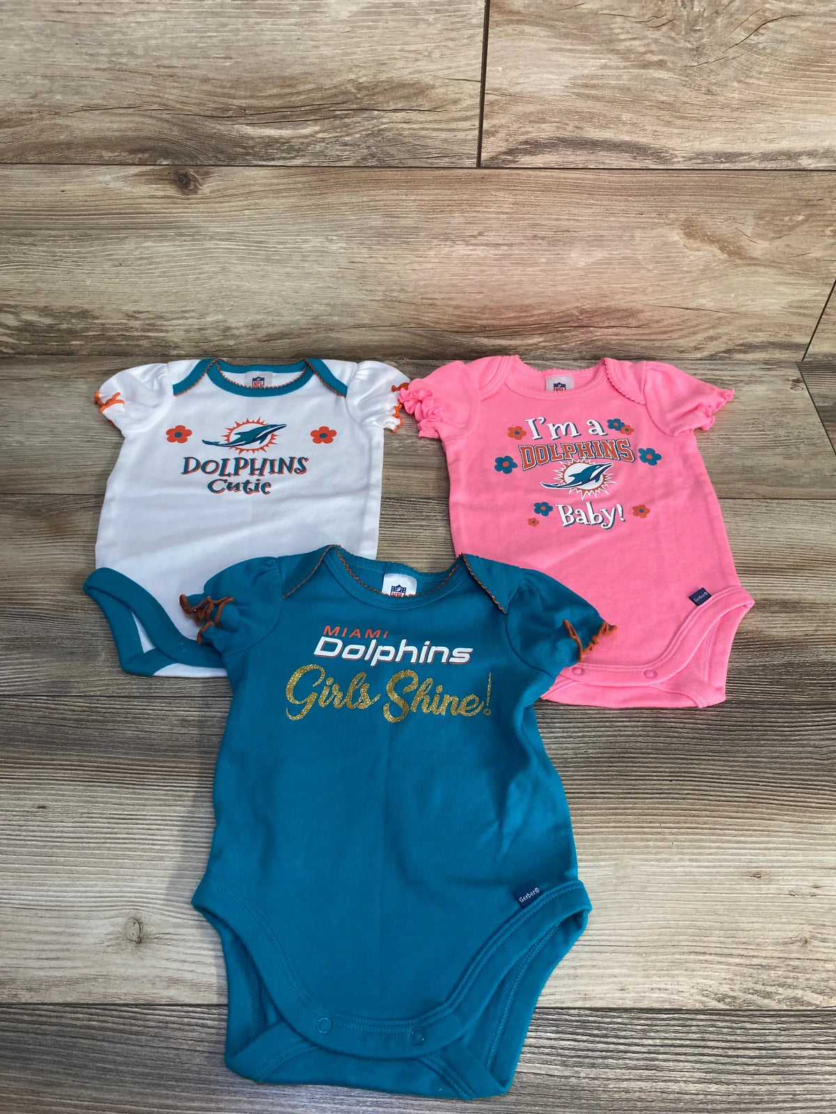 Overhead view of NFL Team Apparel Miami Dolphins three pack bodysuits showcasing three Dolphins graphics on a wood floor