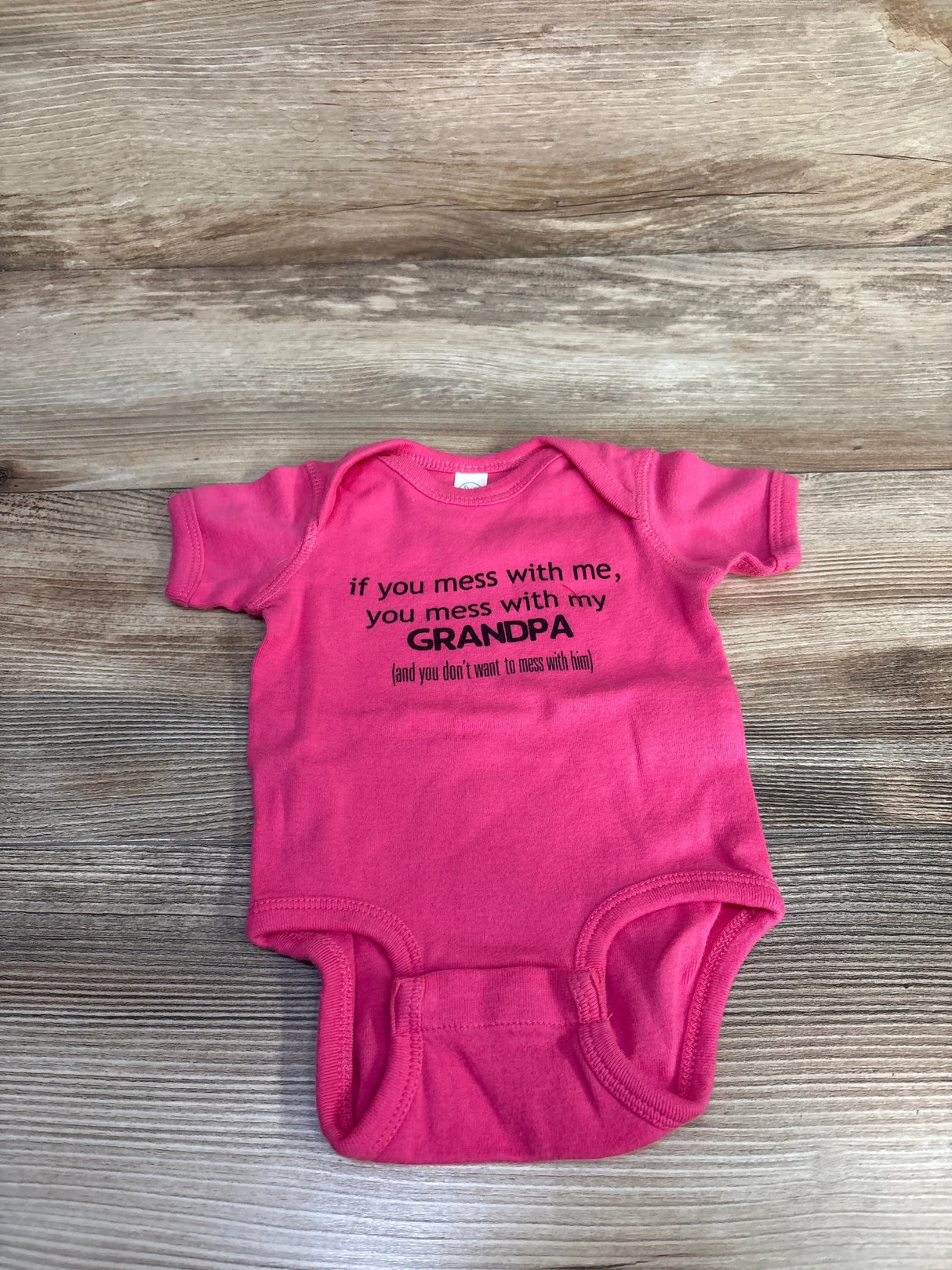 Top-down view of pink 6-month bodysuit with black printed slogan 'if you mess with me, you mess with my GRANDPA' on wood background