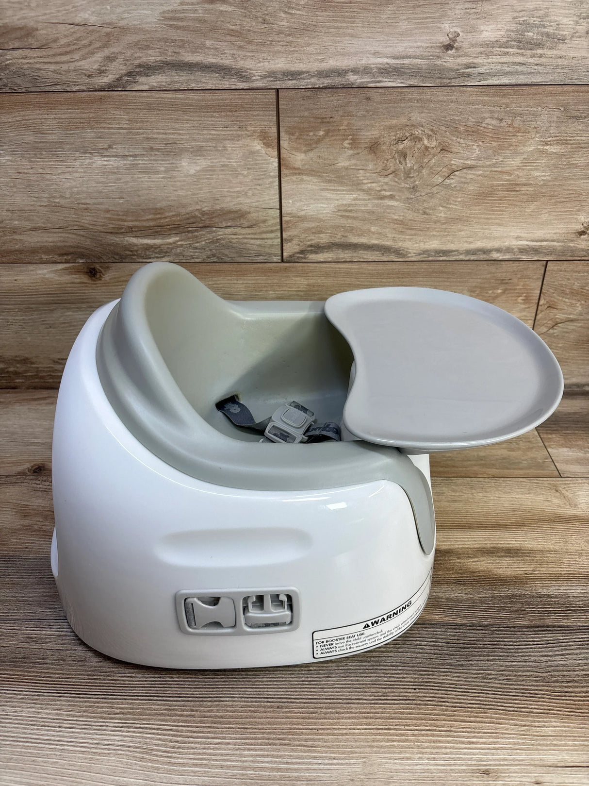 Three-quarter view of Bumbo multi seat (grey insert, white shell) showing removable tray and harness on wood background