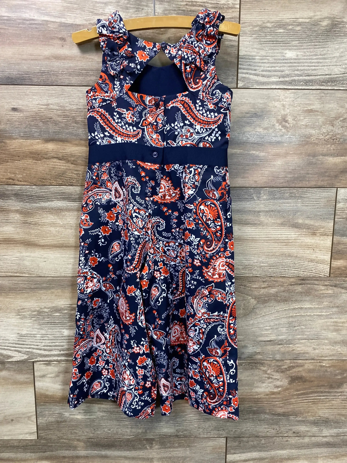 Front view of navy paisley jumpsuit with ruffled straps, keyhole back and buttoned waist, on hanger against wood backdrop
