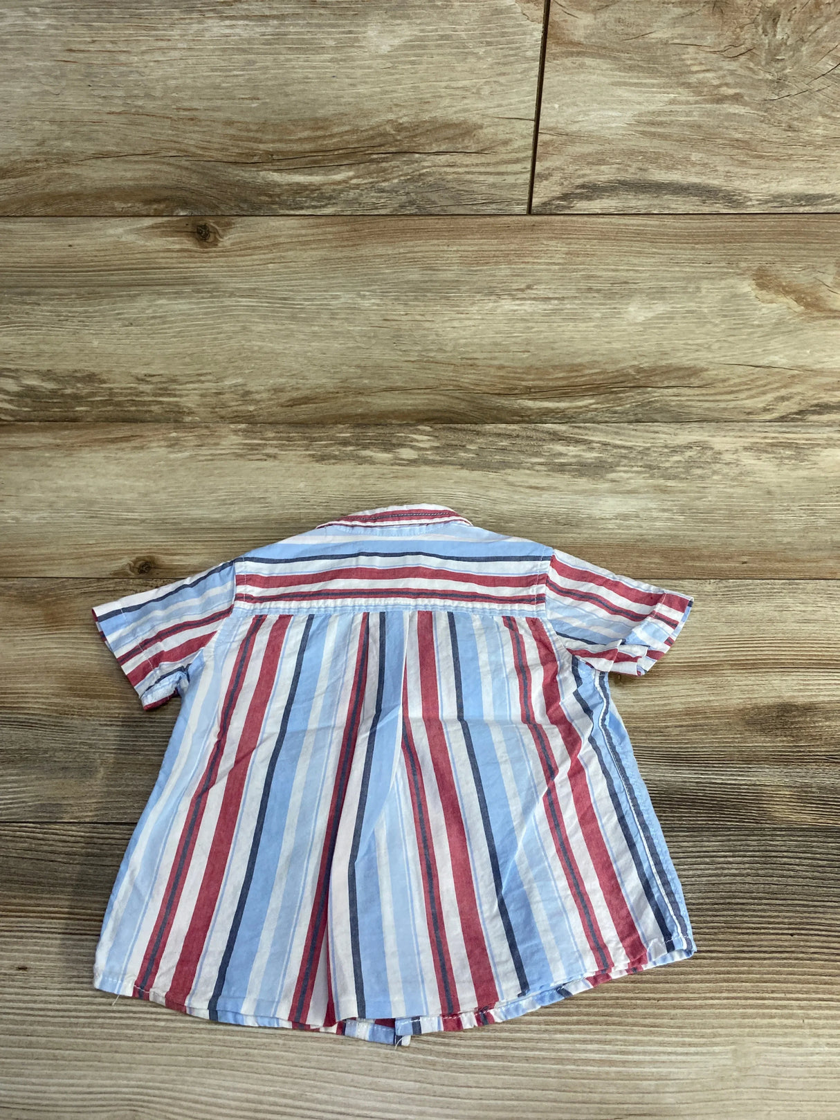 Back view of Childrens Place Striped Button Up Shirt in red white and blue vertical stripes showcasing short sleeves and pleated yoke on wood plank floor