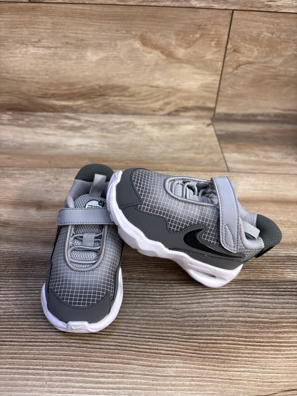 Side view of Nike Air Max Nova TD sneakers in Wolf Grey showcasing hook and loop straps and white cushioning soles on wood