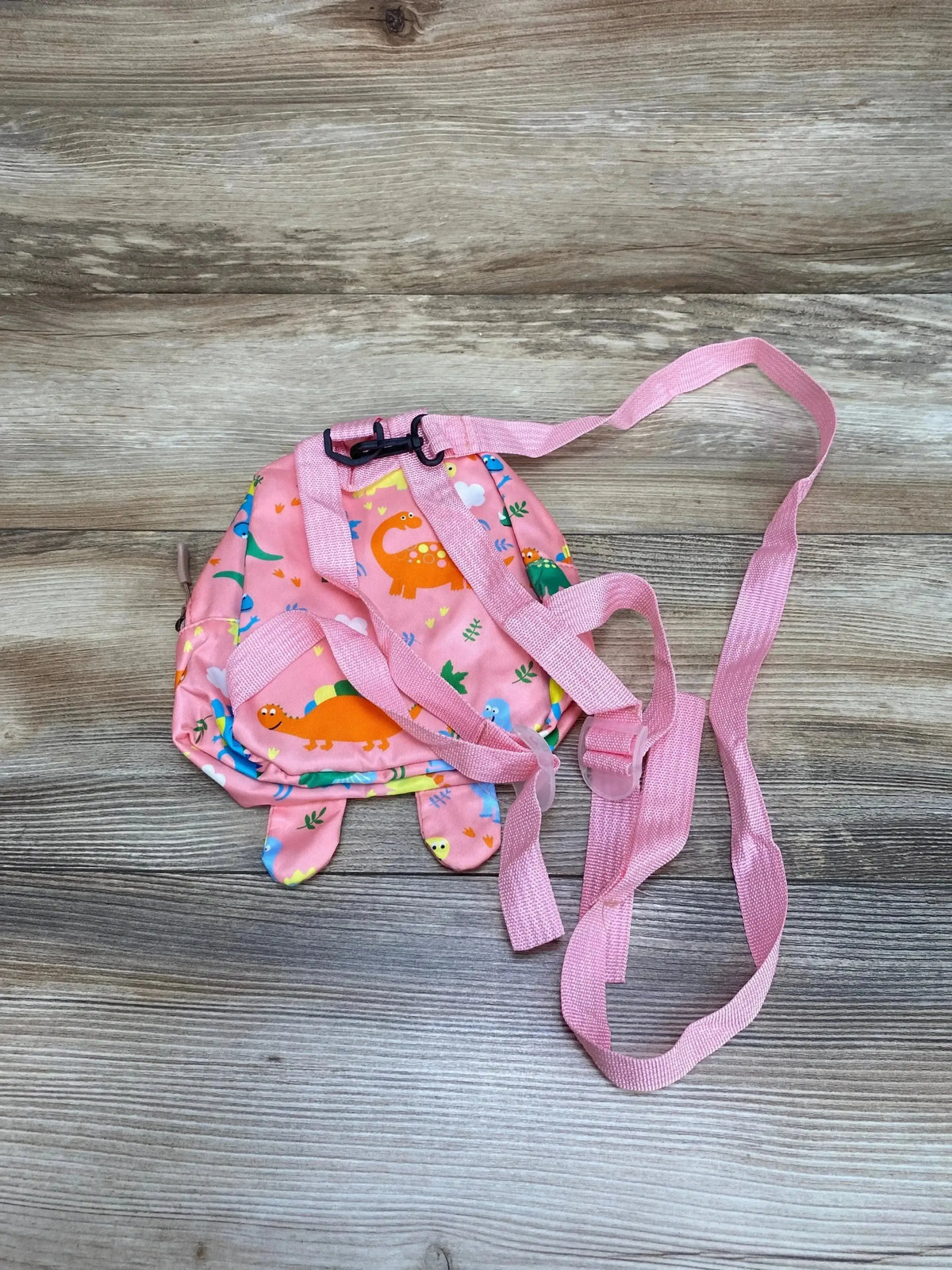 Top-down view of pink dinosaur kids safety harness backpack showing adjustable straps and long leash on wood-plank background