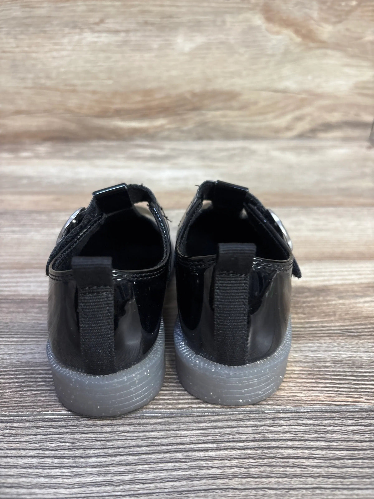 Rear view of Baby Gap T-Strap Loafers showcasing black vegan patent leather uppers and rubber soles on a wood-grain background