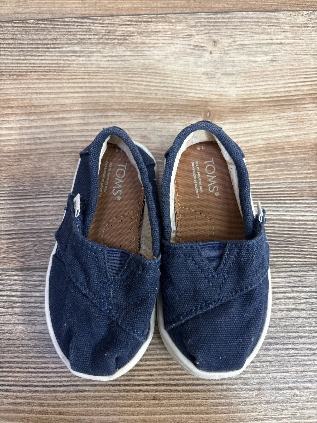 Top-down view of Toms Tiny Alpargata toddler slip-on shoes in navy canvas showing white rubber soles and stitched toe detail on wood background