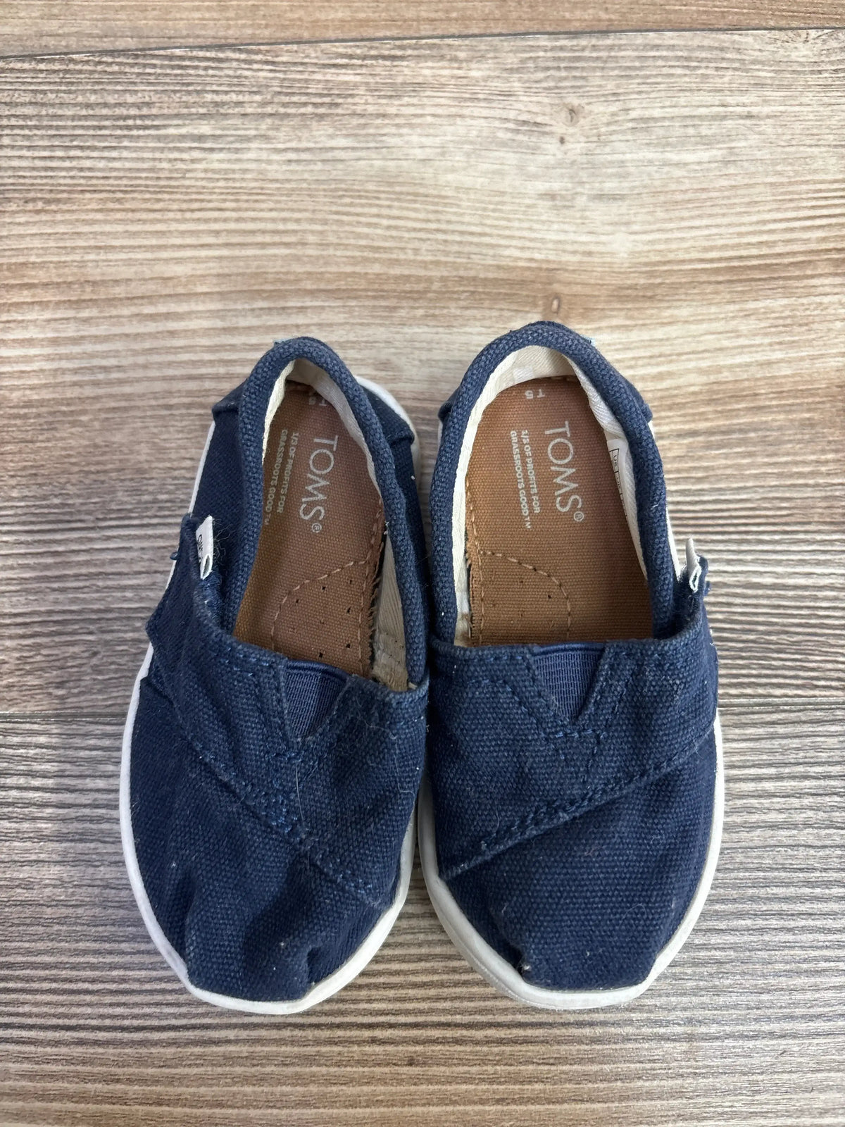 Top-down view of Toms Tiny Alpargata toddler slip-on shoes in navy canvas showing white rubber soles and stitched toe detail on wood background