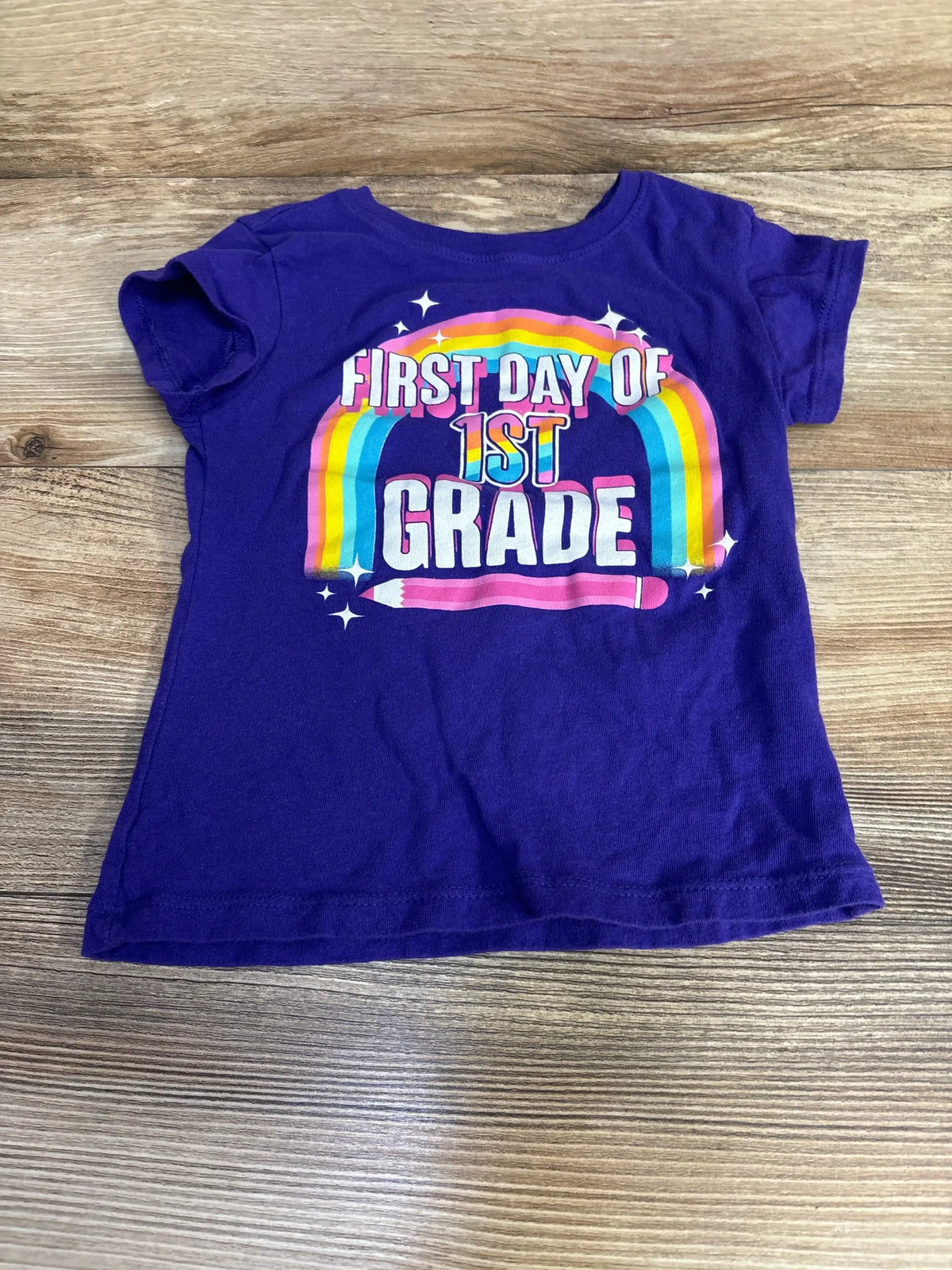 Flat lay of purple Children's Place toddler t-shirt showing FIRST DAY OF 1ST GRADE rainbow and pencil graphic on wooden floor