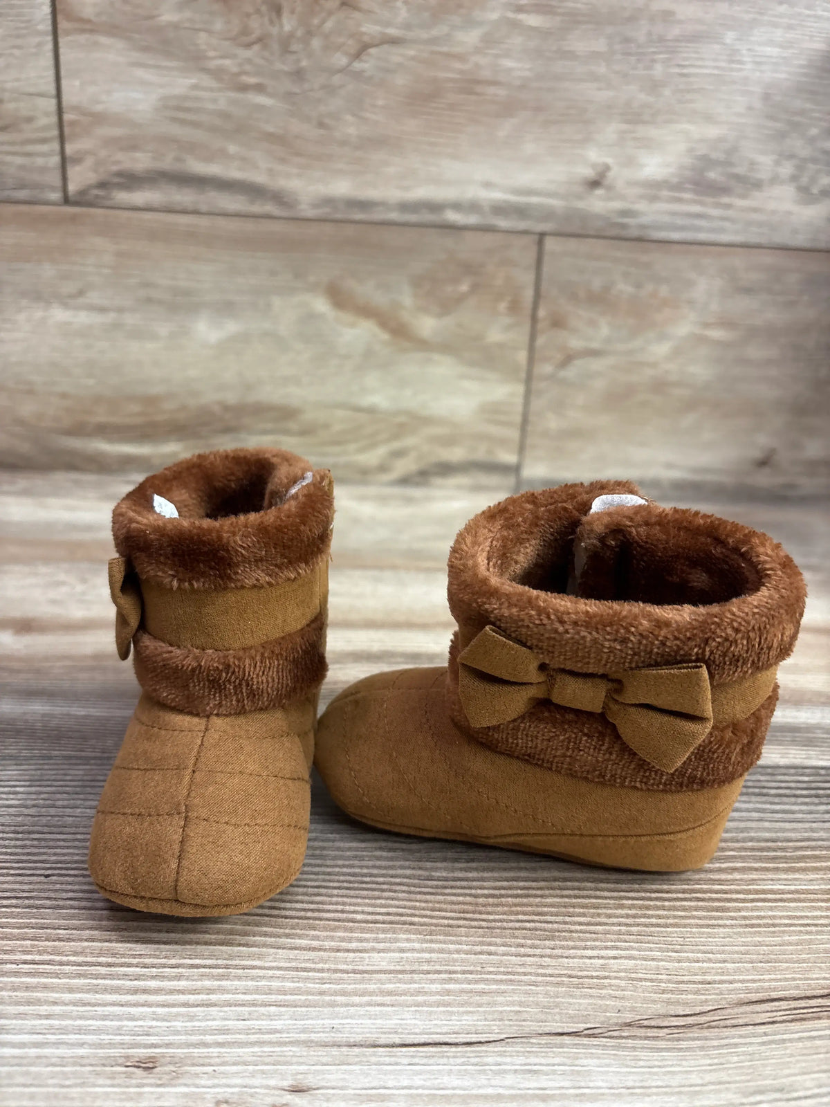 Front and side views of CRYBOKO brown faux fur soft sole boots showcasing bow detail and foldover cuffs on a wood surface