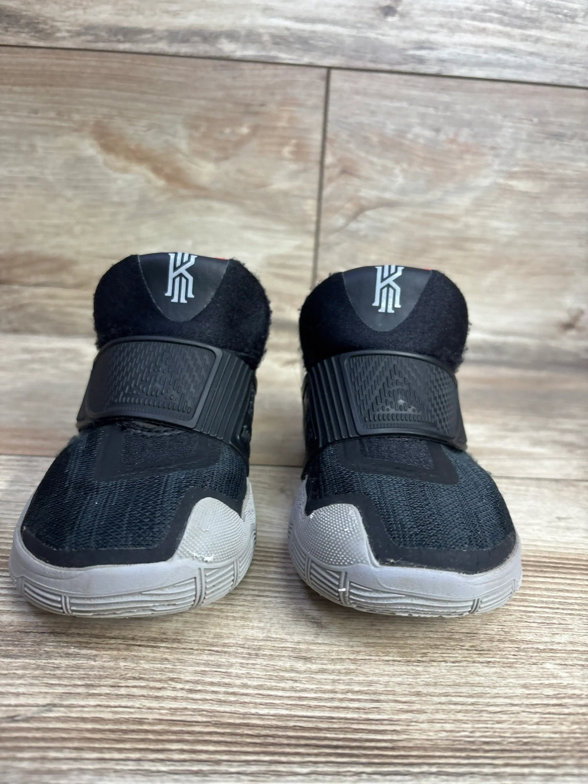 Black and gray toddler athletic shoes with velcro straps and basketball logo, on wood floor