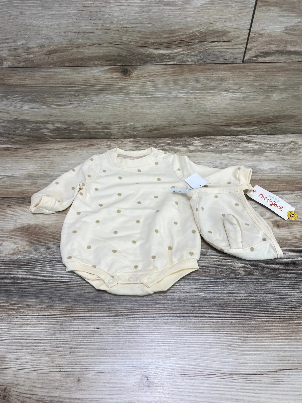 Top-down view of cream Cat & Jack polka-dot baby romper and matching bonnet showing long sleeves and snap crotch on wood background