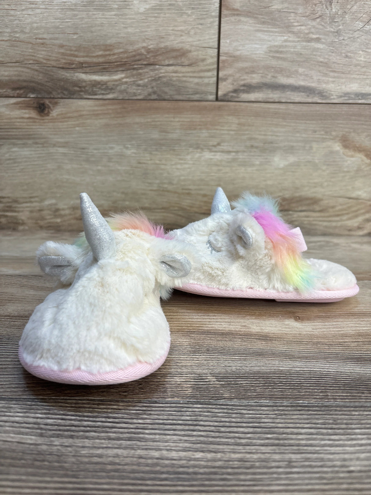 Fluffy unicorn slippers for kids with rainbow mane, silver horn, and pink sole on wood floor