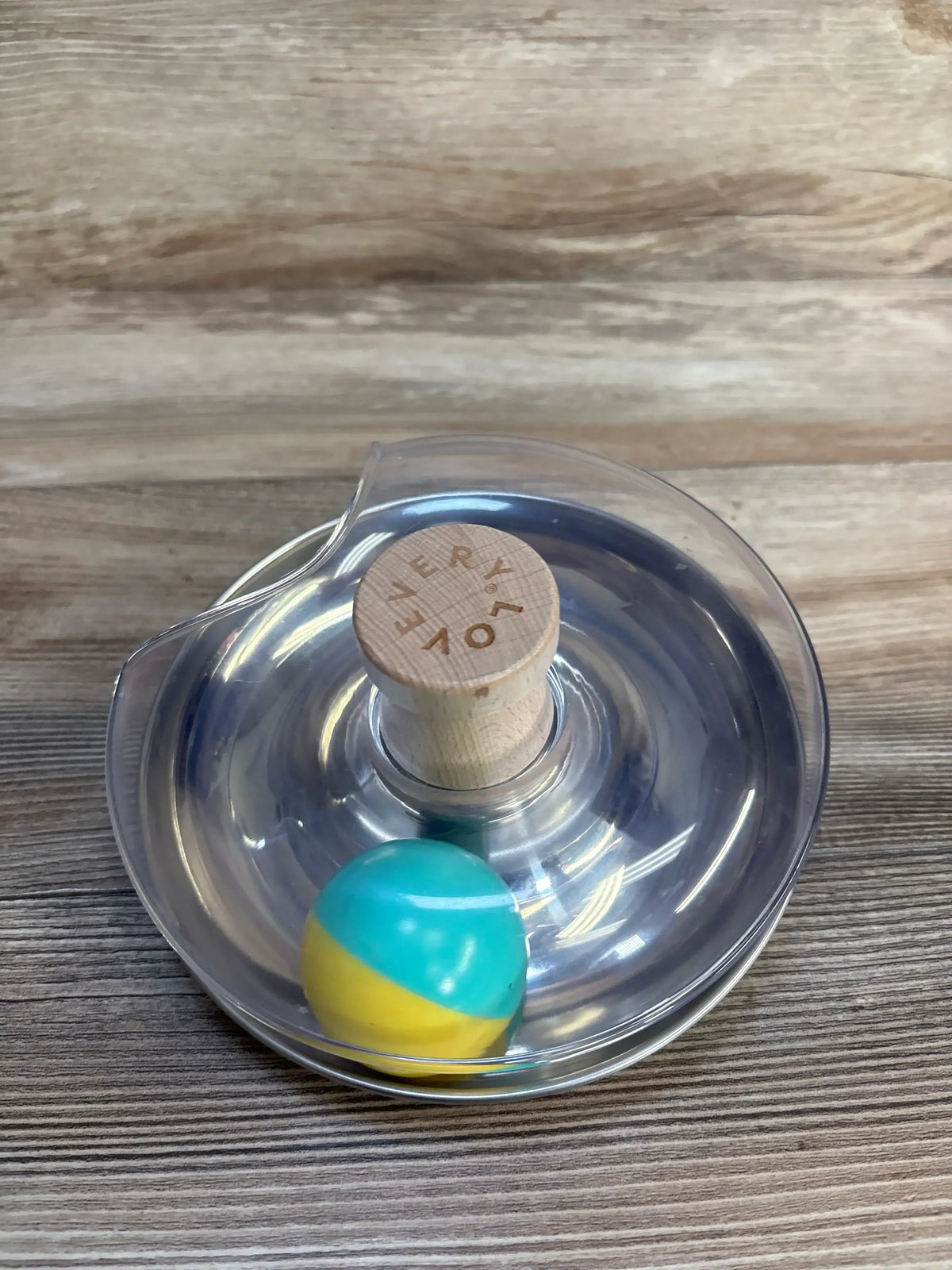 Top-down view of Lovevery Tip & Turn wooden spinner showing engraved LOVEVERY knob and teal-and-yellow ball in clear plastic dish on wood surface