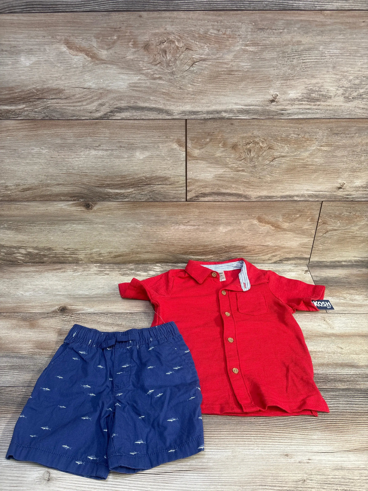 Top-down view of an OshKosh 2-piece set with a red button-up top and navy fish-print woven shorts on a wood floor