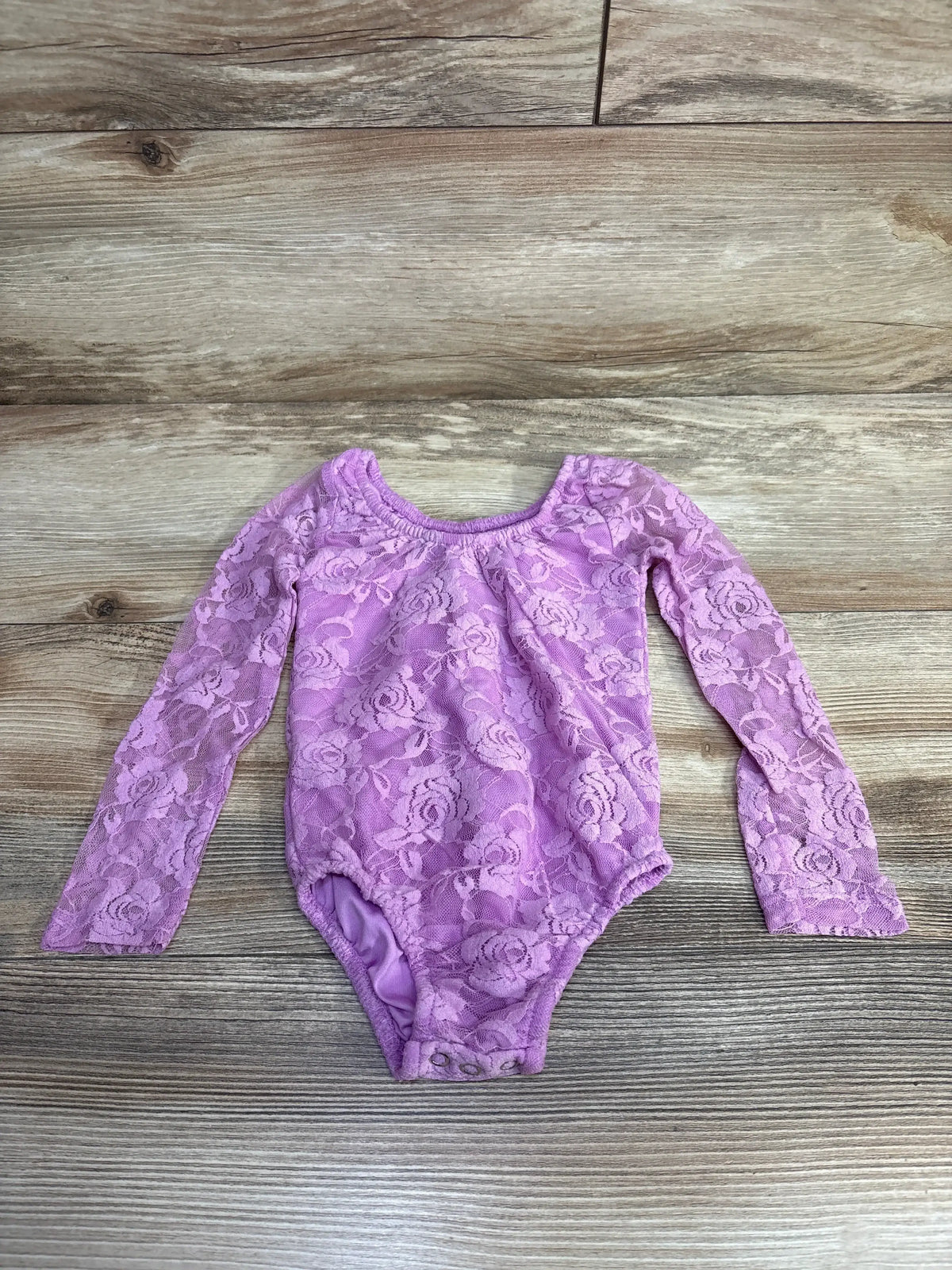 Front view of Bailey’s Blossoms laced long sleeve bodysuit in lilac, size 2T, showcasing floral lace and snap closure on wood