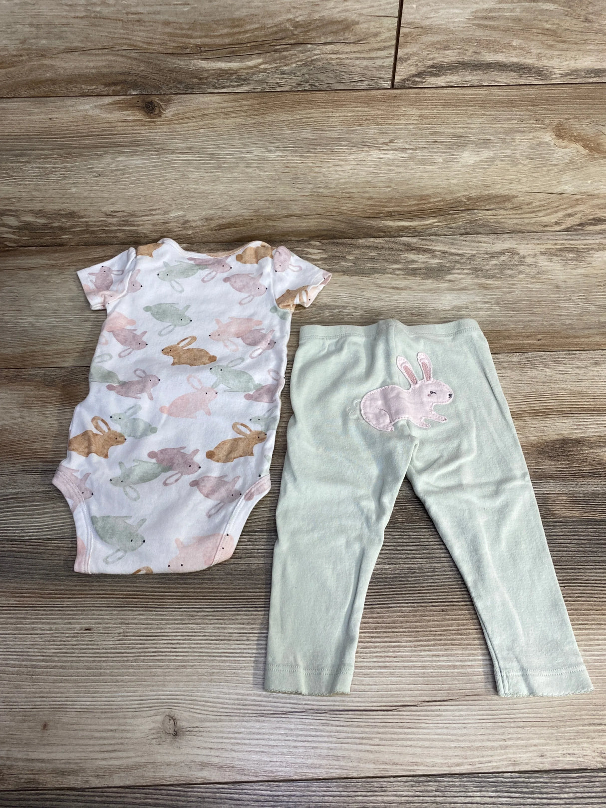 Top-down of Carter's 2-piece baby bodysuit and pants with pastel bunny print and pink bunny patch on mint pants on wood floor