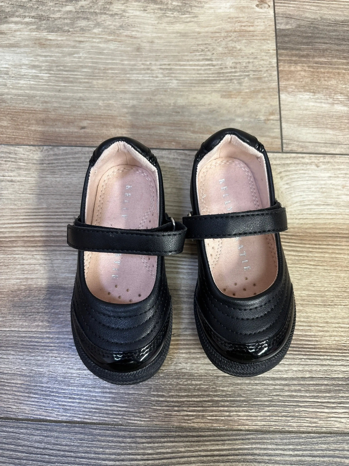 Top view of black Mary Jane toddler flats showing quilted upper, patent toe cap and hook and loop strap on wood floor