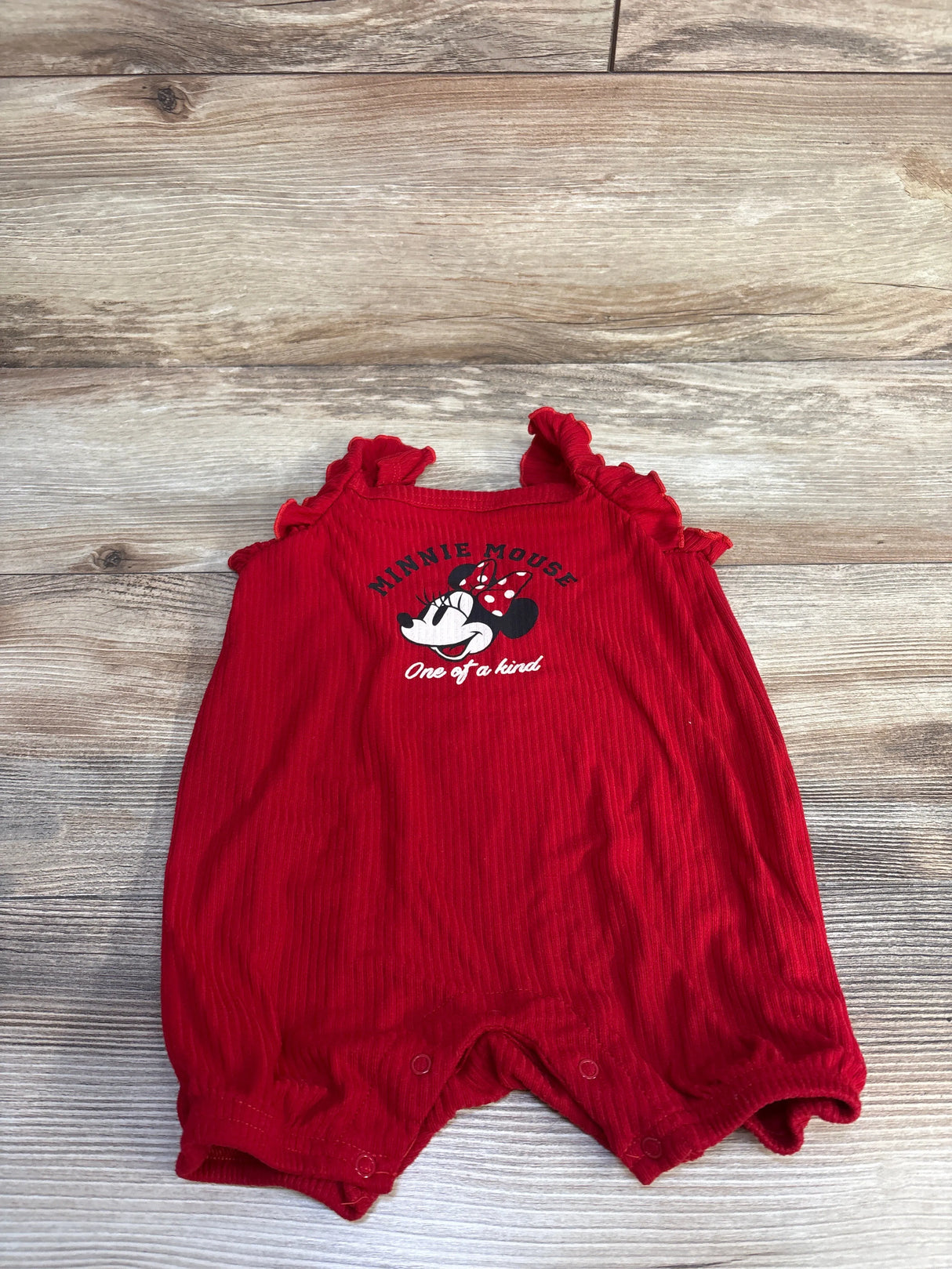 Front view of red Disney Baby shortie romper with Minnie Mouse print, ruffled shoulder straps, snap crotch laid flat on wood background