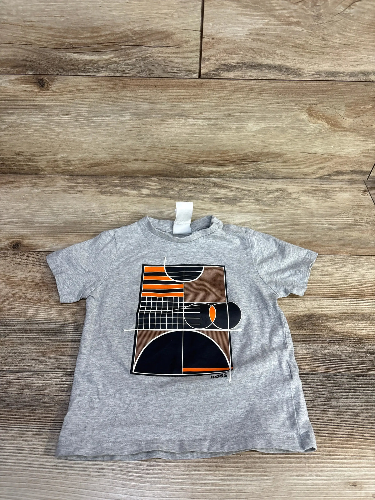 Flat lay of grey Boss Kidswear toddler short-sleeve t-shirt showing geometric orange, black and brown graphic on wood background