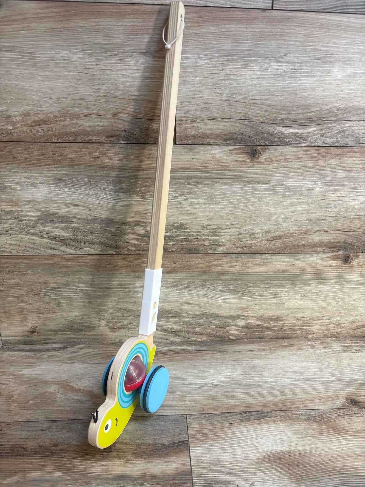 Angled view of Hape Snail Push Pal walking toy showcasing yellow wooden snail head, blue wheels, clear rattle ball, and long wooden push handle on wood floor