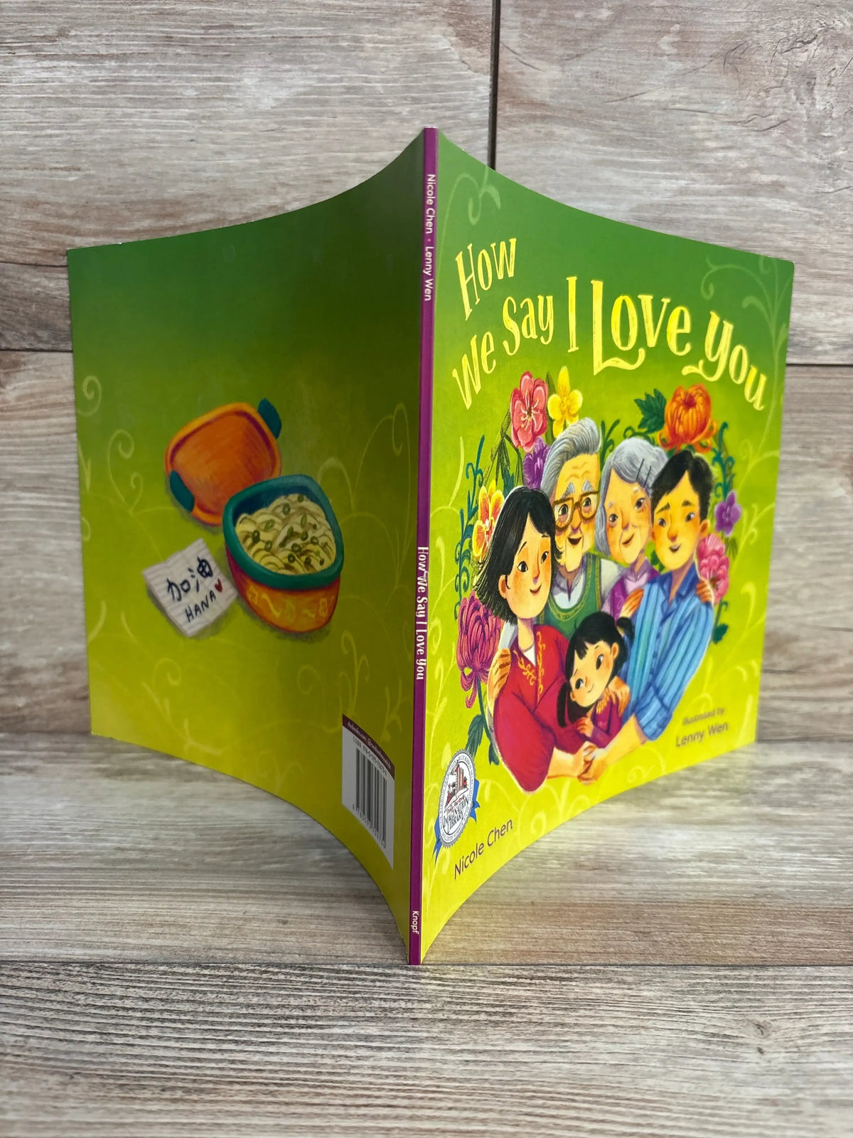 Three-quarter view of How We Say I Love You paperback showing colorful family on front and green back on wood background