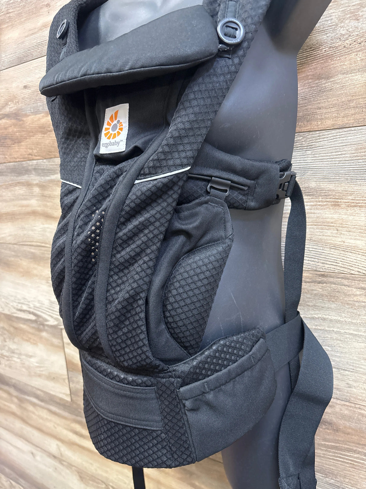 Side view of Ergobaby Omni Breeze baby carrier in black with breathable mesh, waist belt and buckles on wood wall