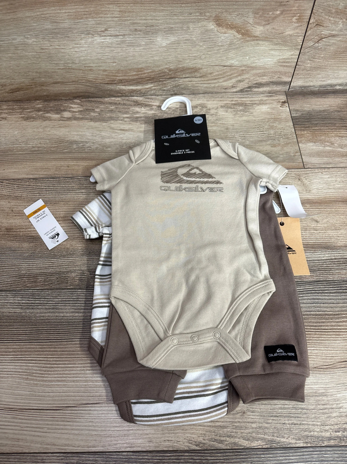 Top-down view of Quiksilver 3-piece newborn set showcasing tan logo bodysuit, striped bodysuit, and brown joggers on wood