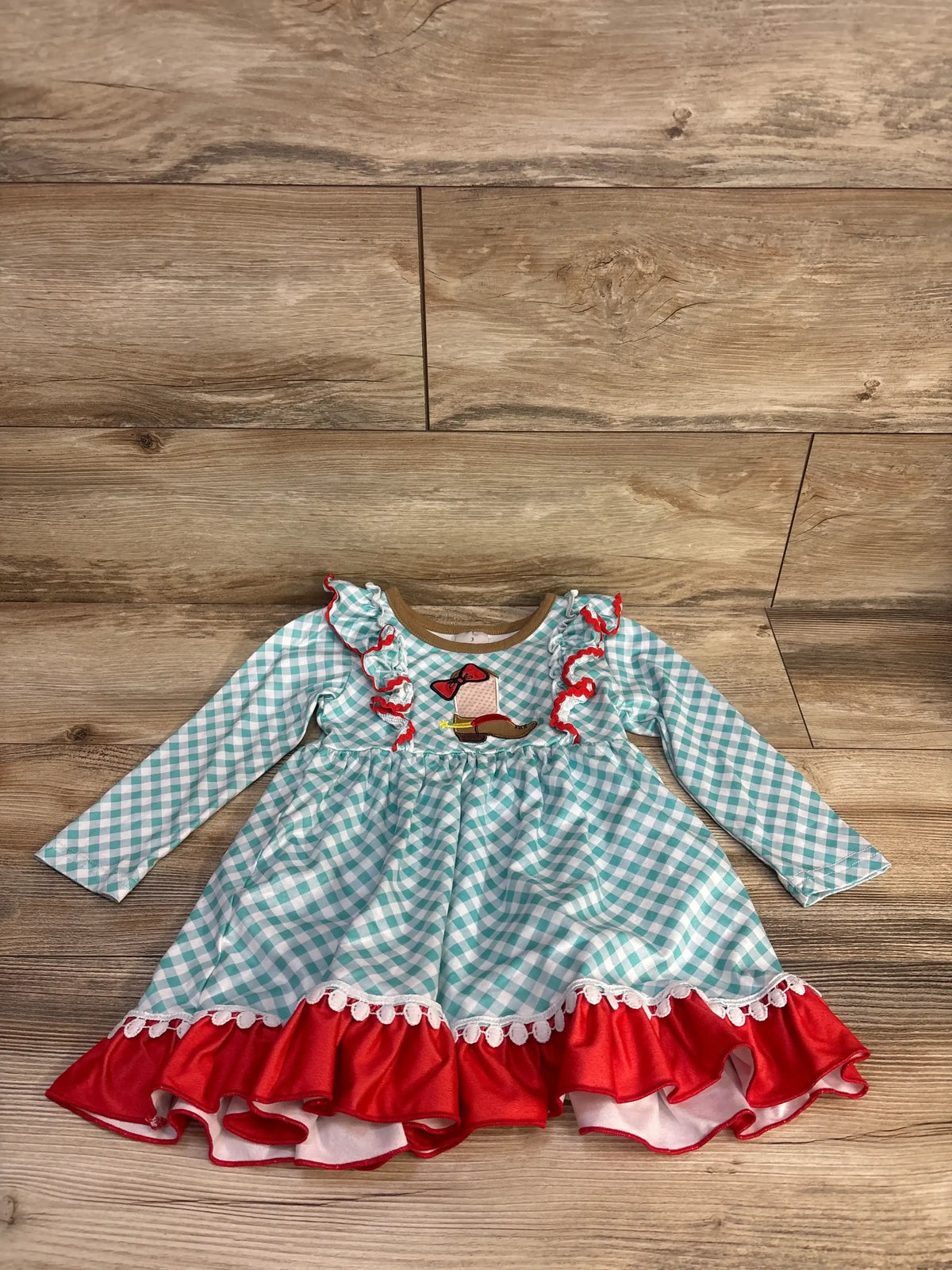 Front flat lay of girls cowgirl boot long sleeve dress size 3T showcasing turquoise gingham pattern, embroidered boot applique and red ruffle hem on wood floor