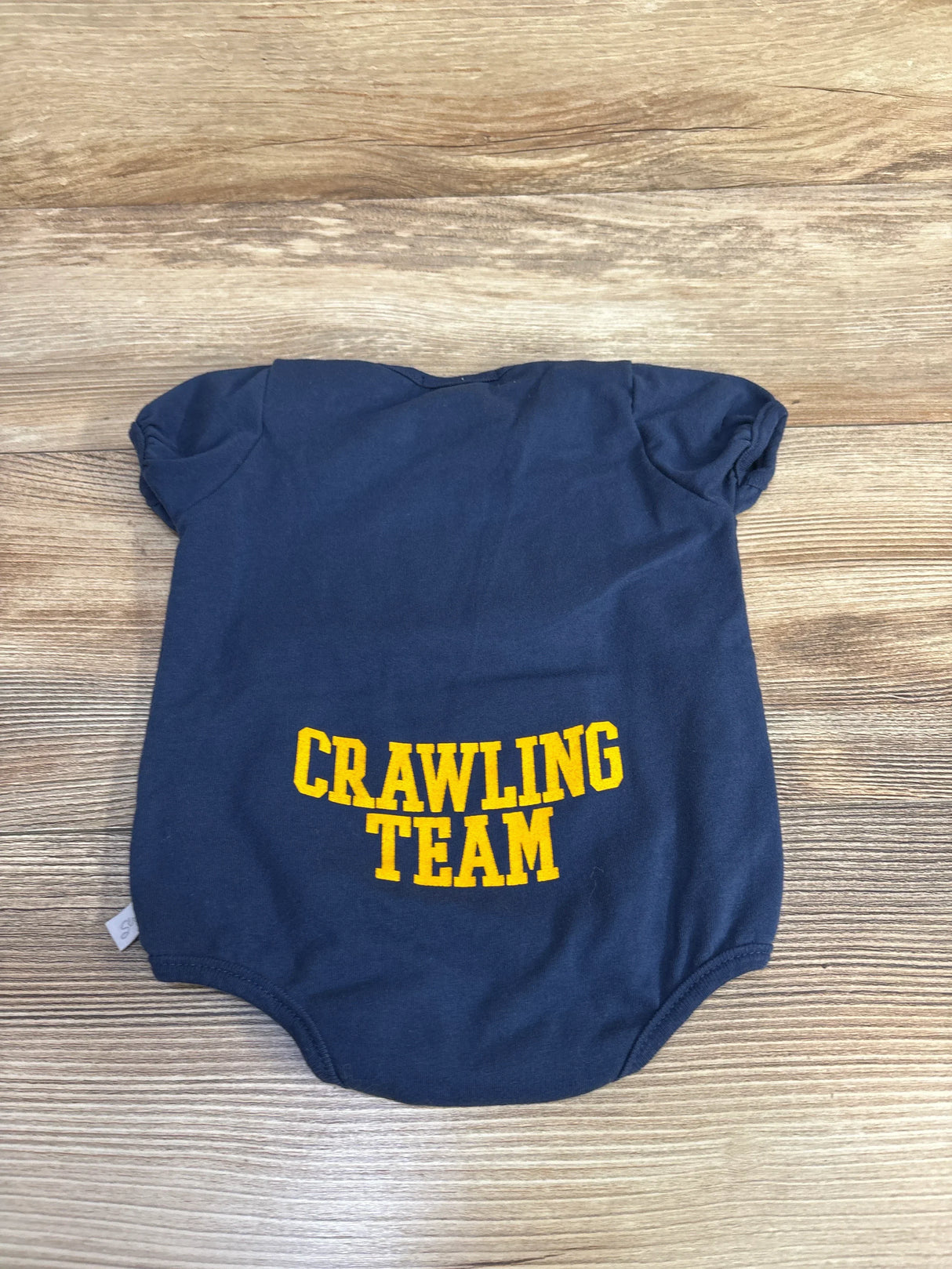 Flat lay of Third Street navy baby bodysuit (3-6 months) showing yellow 'CRAWLING TEAM' text on wood plank background