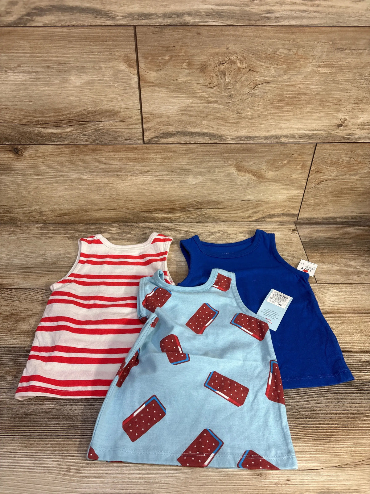 Front view of three Cat & Jack sleeveless ice cream tank tops in blue, light blue, and red-striped designs on a wood floor